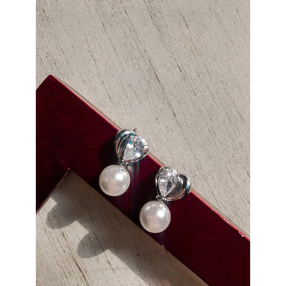Two elegant silver drop earrings, each featuring a white pearl hanging below a silver heart with a clear, pear-shaped stone, displayed on a textured surface.