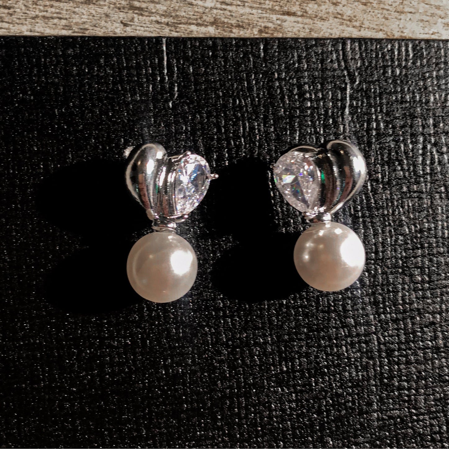 Two elegant silver drop earrings, each featuring a white pearl hanging below a silver heart with a clear, pear-shaped stone, displayed on a textured surface.