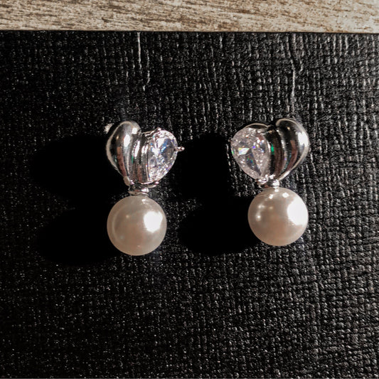 Two elegant silver drop earrings, each featuring a white pearl hanging below a silver heart with a clear, pear-shaped stone, displayed on a textured surface.