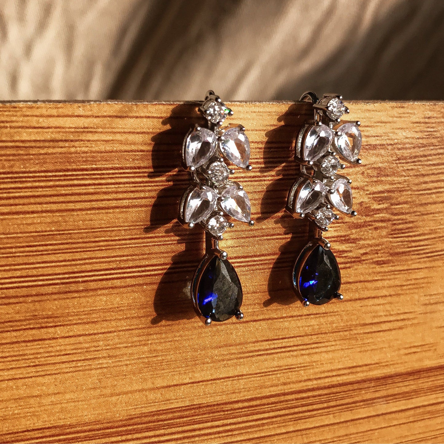 Two silver dangle earrings, one with a black teardrop crystal and the other with a blue teardrop crystal, both featuring clear sparkling stones above the teardrop, displayed on a wooden surface.