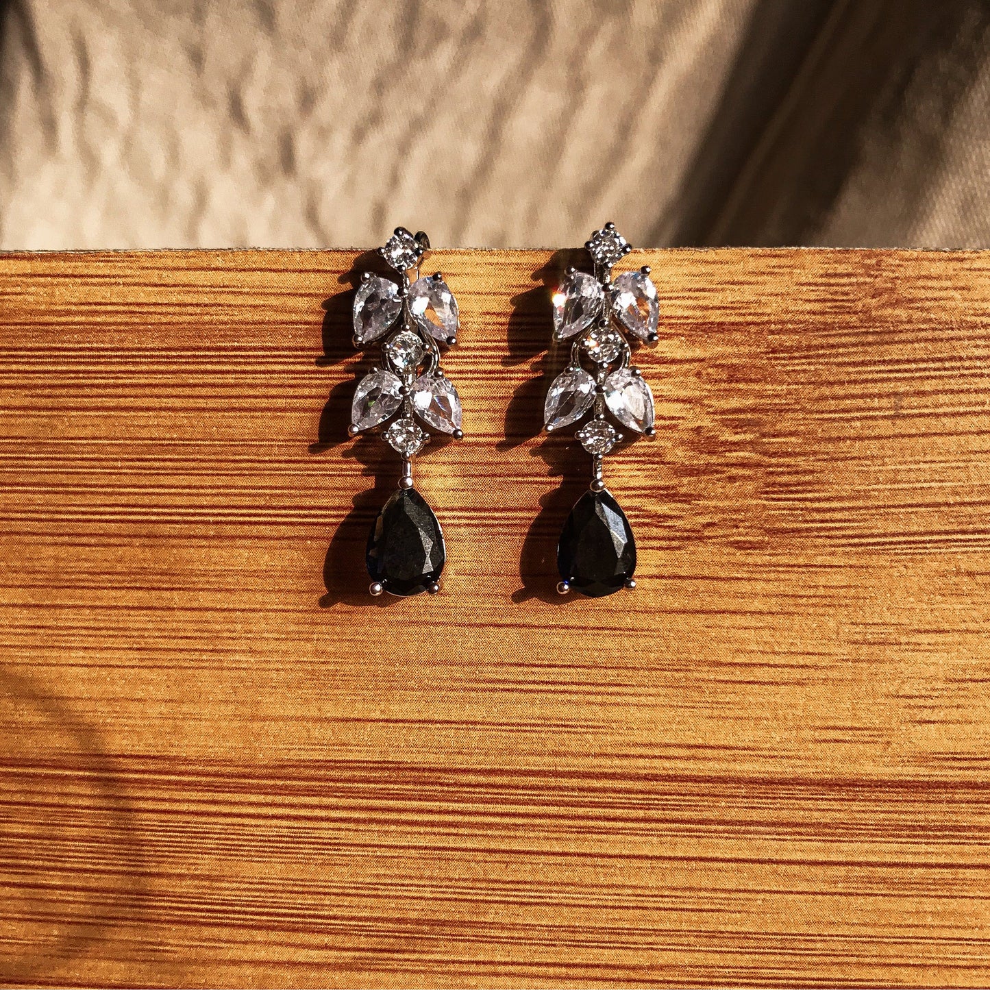 Two silver dangle earrings, one with a black teardrop crystal and the other with a blue teardrop crystal, both featuring clear sparkling stones above the teardrop, displayed on a wooden surface.