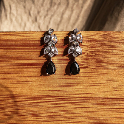 Two silver dangle earrings, one with a black teardrop crystal and the other with a blue teardrop crystal, both featuring clear sparkling stones above the teardrop, displayed on a wooden surface.