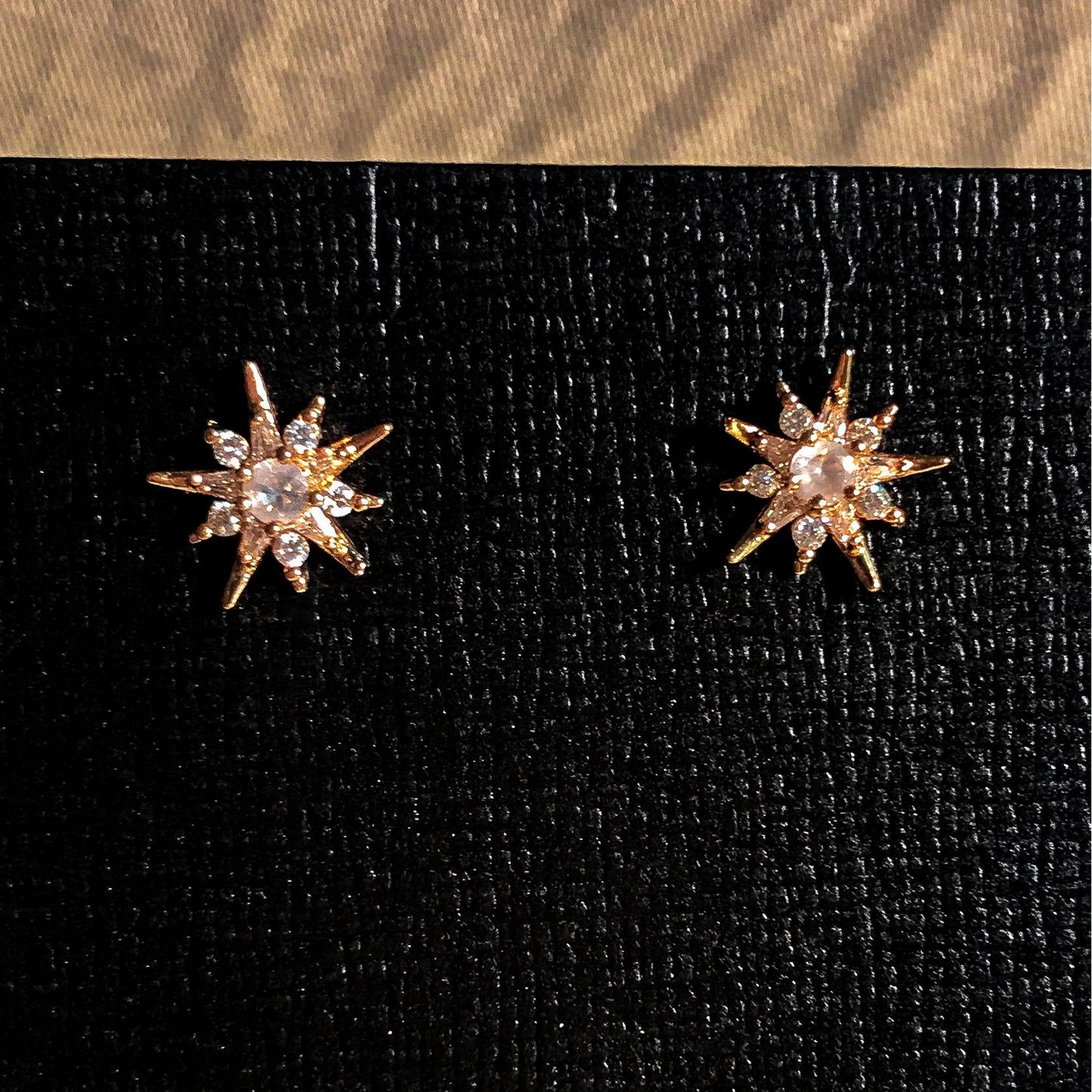 Close-up of two rose gold starburst stud earrings with clear, sparkling stones in the center, displayed on a textured black earring card.