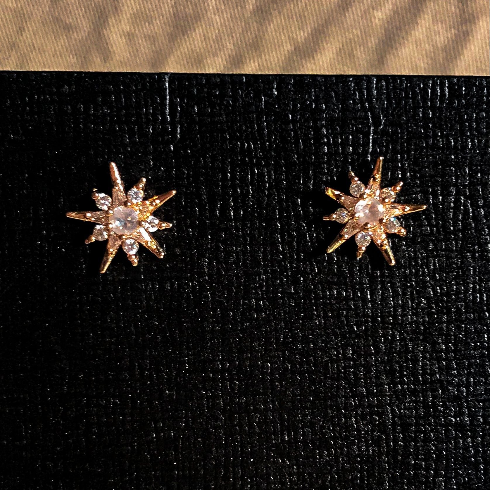 Close-up of two rose gold starburst stud earrings with clear, sparkling stones in the center, displayed on a textured black earring card.
