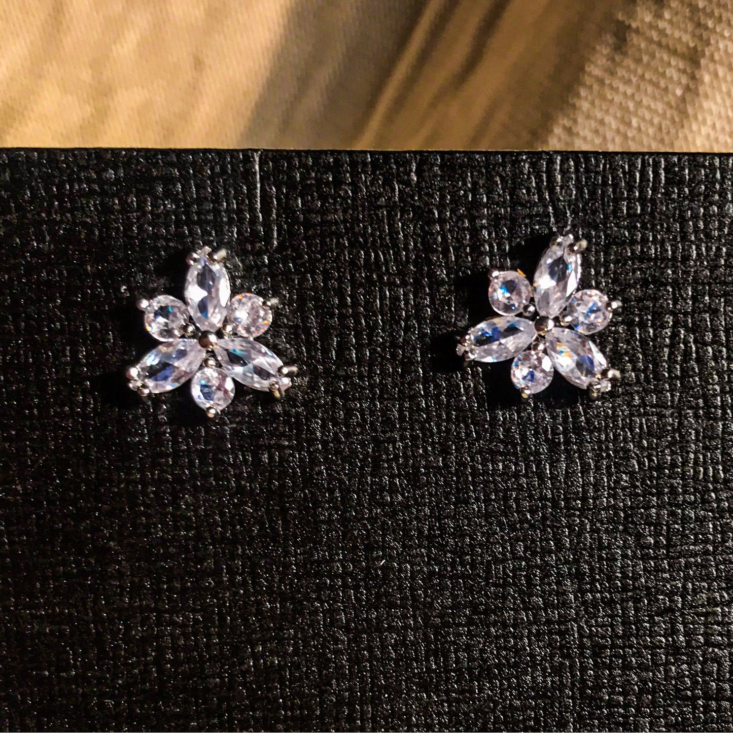 Two small, silver flower-shaped stud earrings set with clear, sparkling stones, displayed on a black earring card.