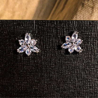 Two small, silver flower-shaped stud earrings set with clear, sparkling stones, displayed on a black earring card.