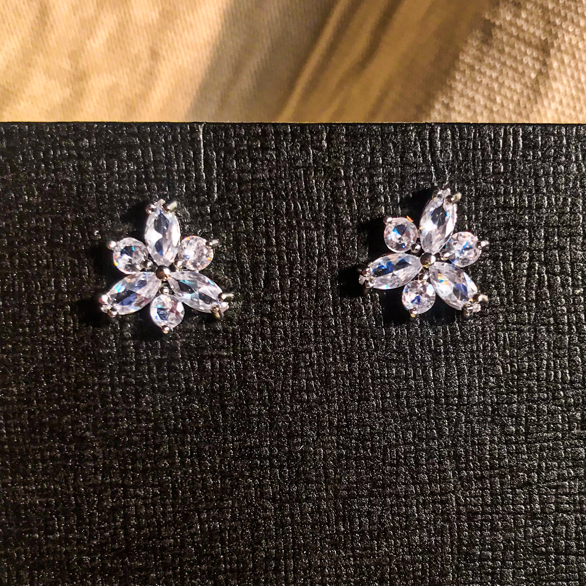 Two small, silver flower-shaped stud earrings set with clear, sparkling stones, displayed on a black earring card.