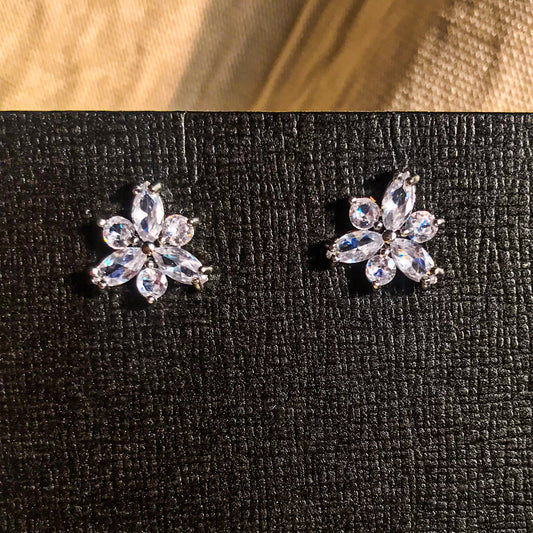 Two small, silver flower-shaped stud earrings set with clear, sparkling stones, displayed on a black earring card.