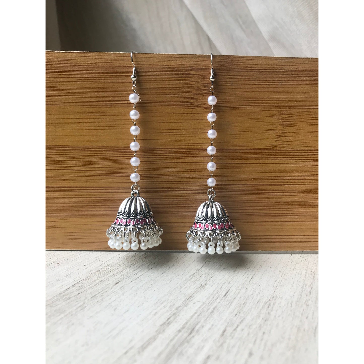 Pair of silver jhumka earrings with pink accents and pearl beaded chains hanging on a wooden backdrop against a white surface