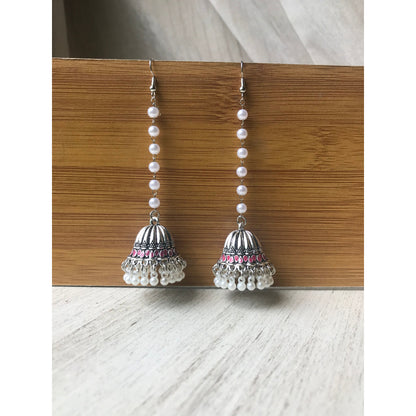 Pair of silver jhumka earrings with pink accents and pearl beaded chains hanging on a wooden backdrop against a white surface
