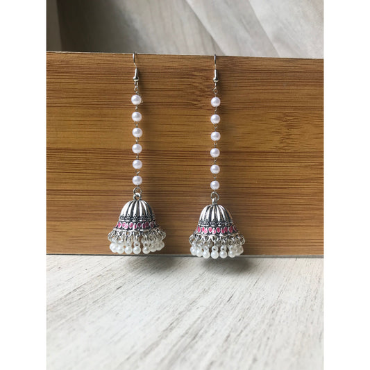 Pair of silver jhumka earrings with pink accents and pearl beaded chains hanging on a wooden backdrop against a white surface