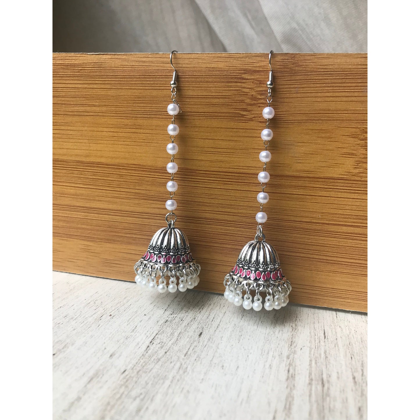 Pair of silver jhumka earrings with pink accents and pearl beaded chains hanging on a wooden backdrop against a white surface