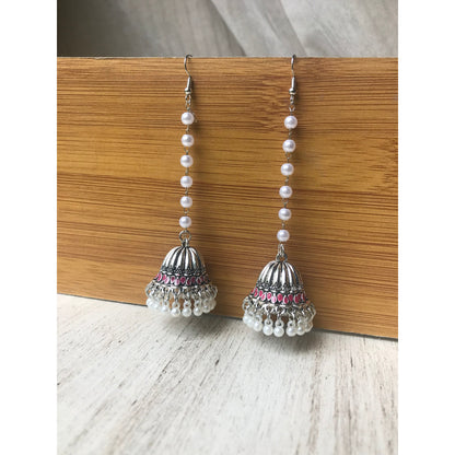 Pair of silver jhumka earrings with pink accents and pearl beaded chains hanging on a wooden backdrop against a white surface