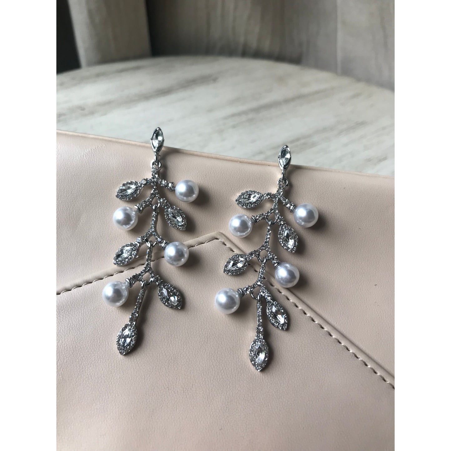 Pair of sparkling crystal leaf dangle earrings displayed on a gray jewelry case with a wood grain surface & curtain backdrop