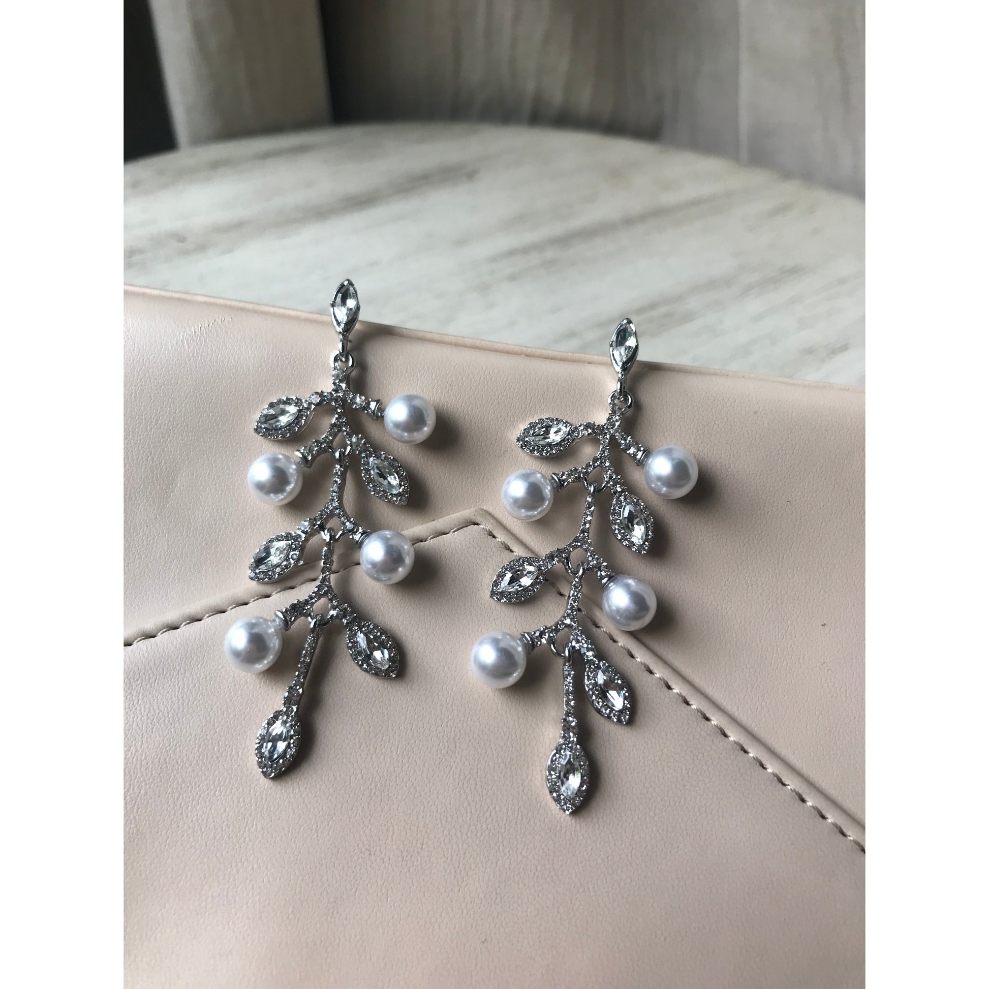 Pair of sparkling crystal leaf dangle earrings displayed on a gray jewelry case with a wood grain surface & curtain backdrop