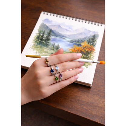 Woman wearing multi-colour twist solitaire silver ring collection while holding paintbrush over sketchbook painting