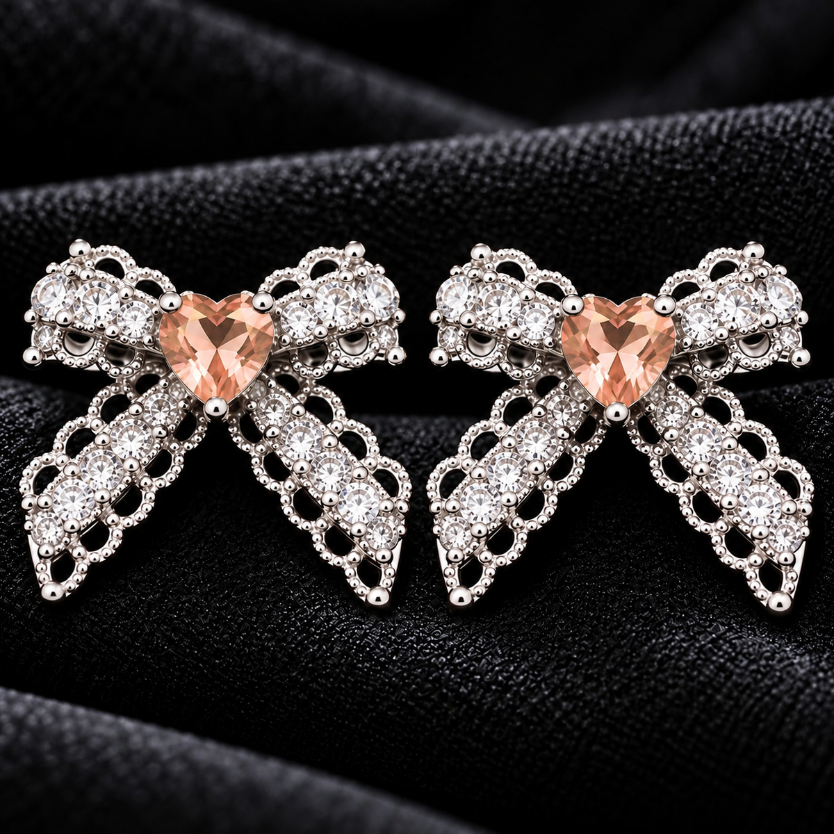 Close-up of Amorette Bow Studs featuring champagne heart-shaped centre stones, clear zircon detailing, and polished silver-tone bow designs on black fabric.