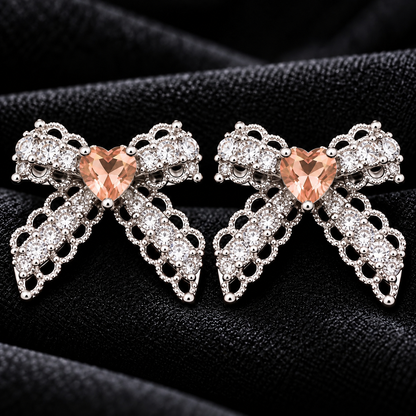 Close-up of Amorette Bow Studs featuring champagne heart-shaped centre stones, clear zircon detailing, and polished silver-tone bow designs on black fabric.
