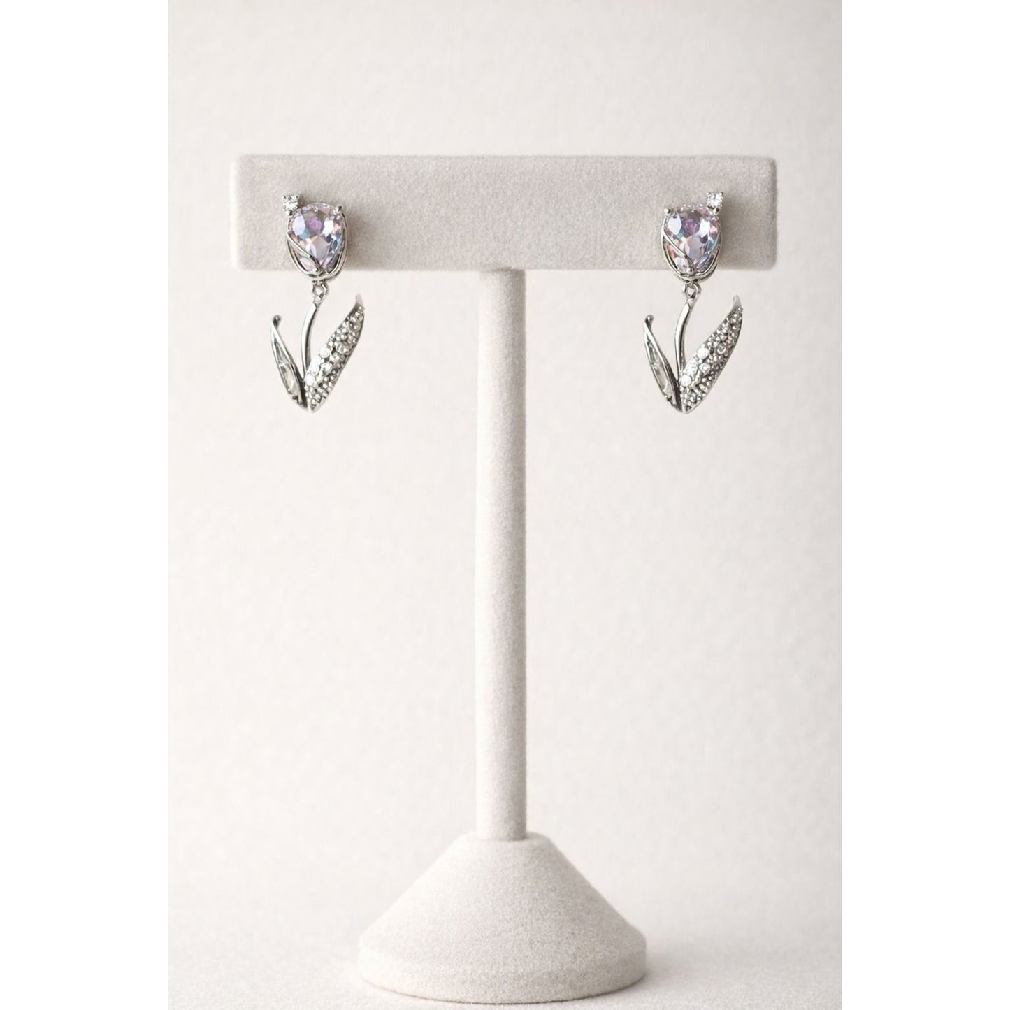 Pair of silver tulip-inspired drop earrings with soft lavender crystal  stones displayed on a white velvet T-shaped jewelry stand against a neutral background