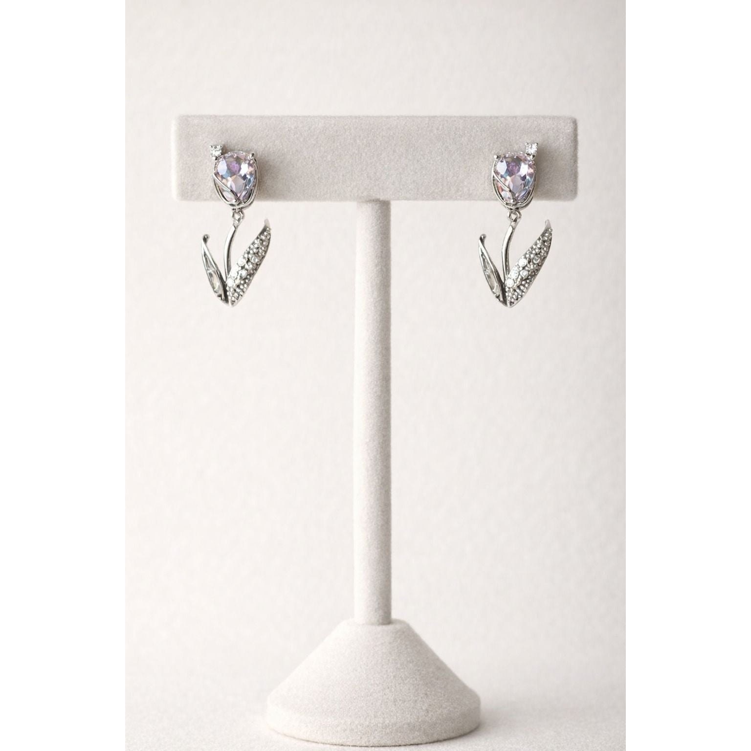 Pair of silver tulip-inspired drop earrings with soft lavender crystal  stones displayed on a white velvet T-shaped jewelry stand against a neutral background