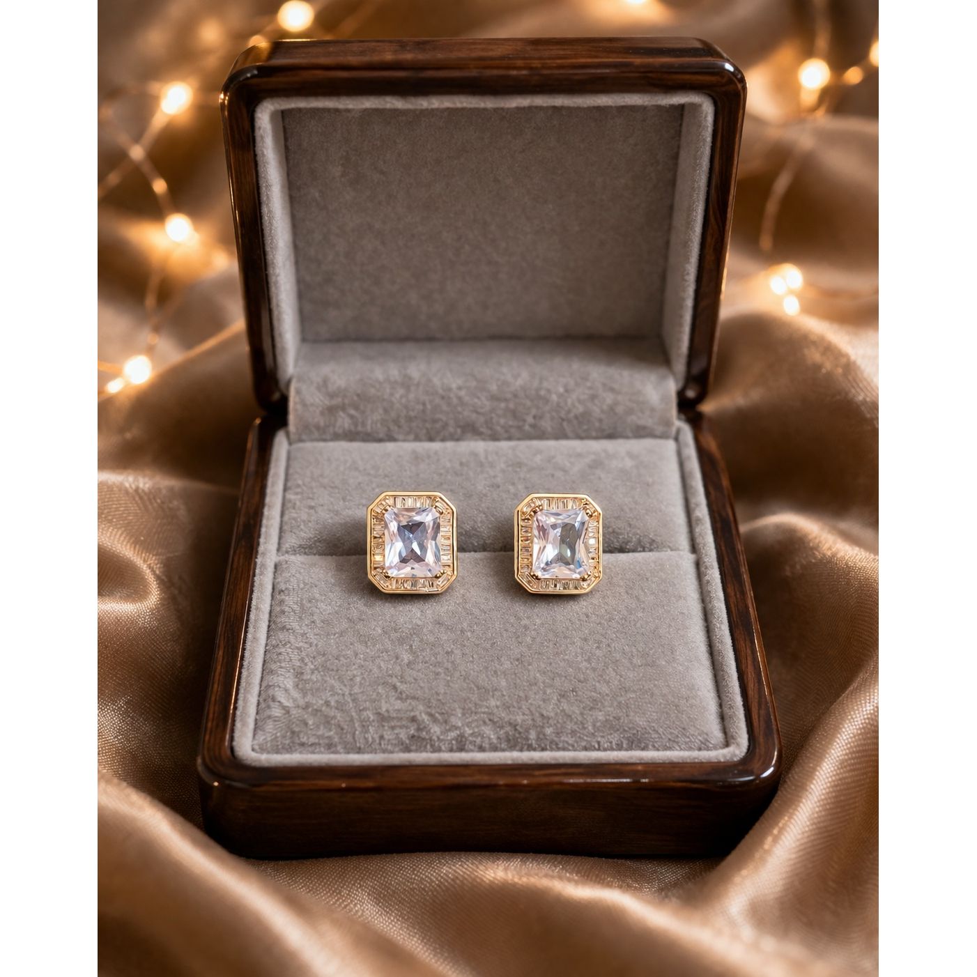 Gold and silver emerald-cut crystal stud earrings displayed in premium jewelry box on satin fabric with warm lights