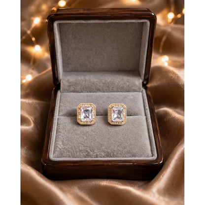 Gold and silver emerald-cut crystal stud earrings displayed in premium jewelry box on satin fabric with warm lights