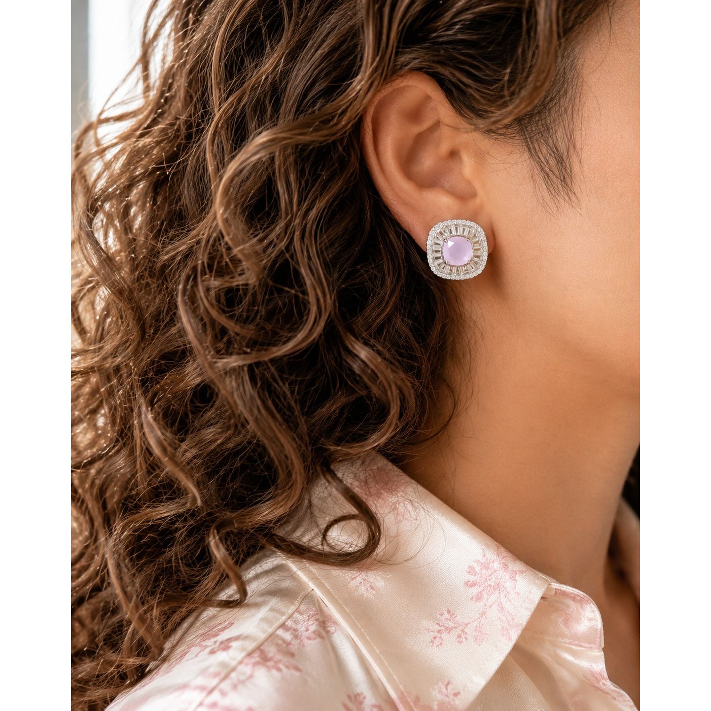 Woman wearing silver halo stud earrings with soft blush pink center stone and crystal baguette frame, paired with a blush floral satin shirt and soft curls.