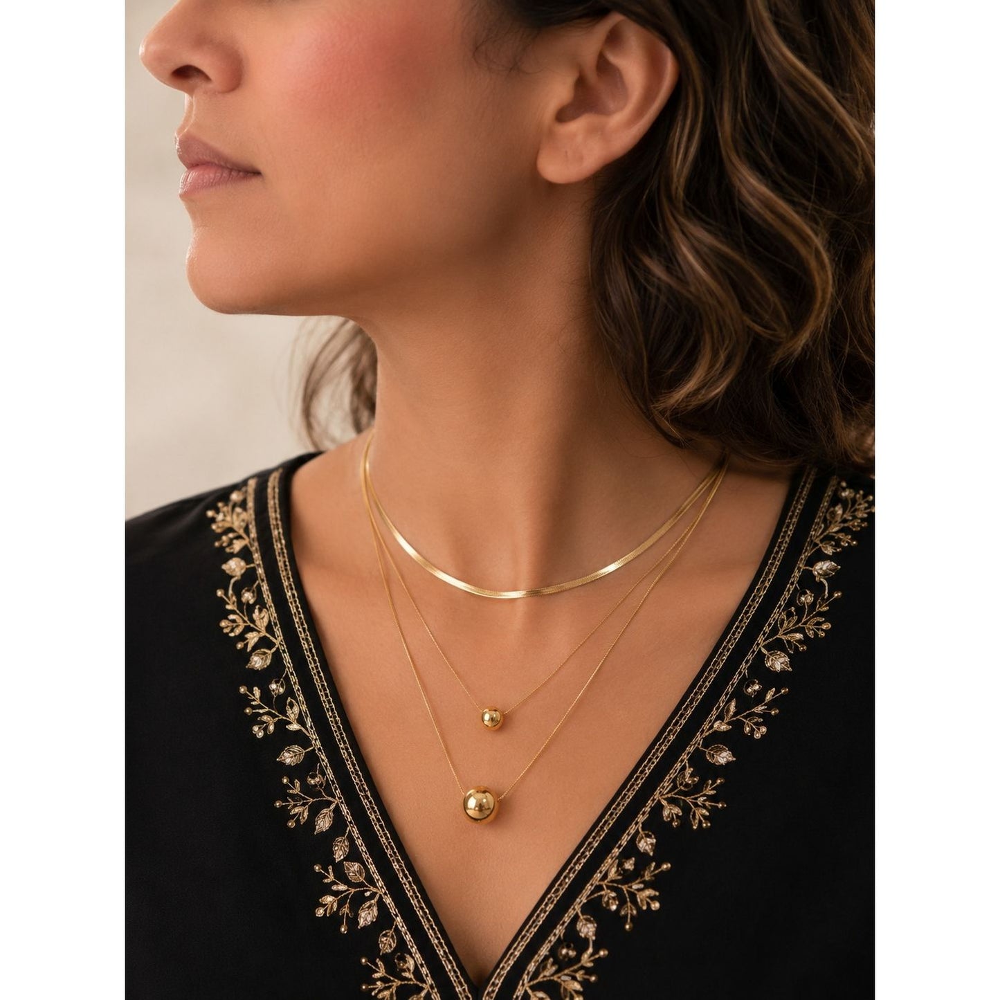 Woman wearing a layered gold necklace featuring polished sphere pendants and a herringbone chain with a black embroidered outfit.