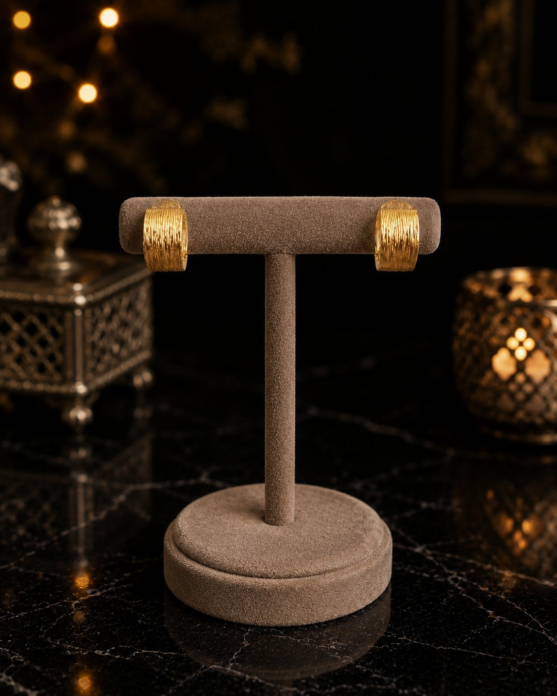 Pair of textured gold hoop earrings displayed on a premium velvet jewelry stand against a luxury black marble background with warm ambient lighting.