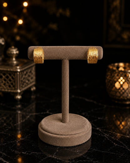 Pair of textured gold hoop earrings displayed on a premium velvet jewelry stand against a luxury black marble background with warm ambient lighting.
