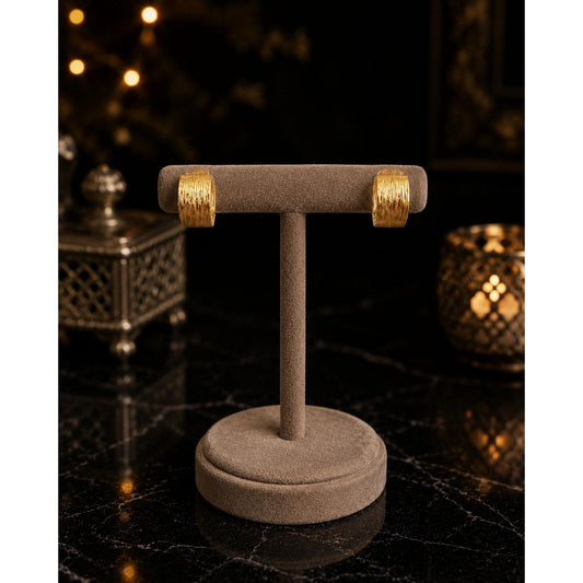 Pair of textured gold hoop earrings displayed on a premium velvet jewelry stand against a luxury black marble background with warm ambient lighting.
