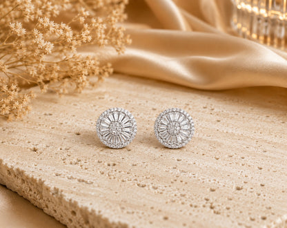 Round silver crystal stud earrings featuring baguette-cut and round-cut stones displayed on a premium travertine stone slab with warm luxury lighting and champagne satin backdrop.