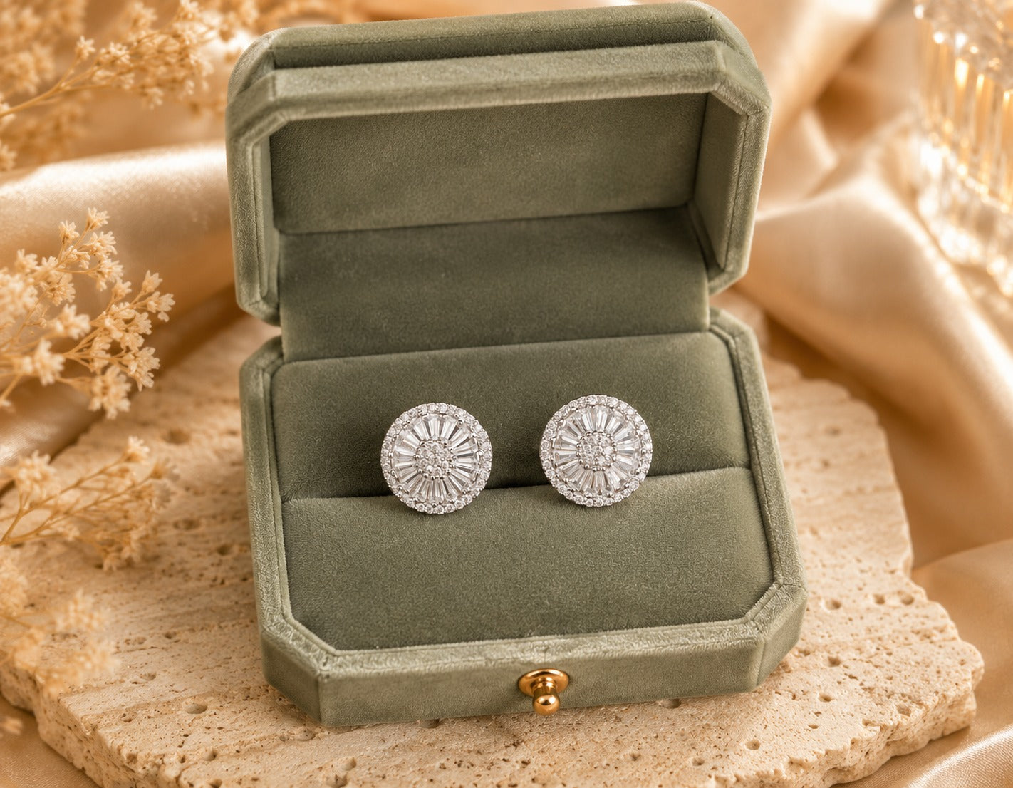 Silver-tone halo crystal stud earrings displayed inside a premium sage green velvet jewelry box on a natural travertine stone surface with champagne satin and dried floral accents.