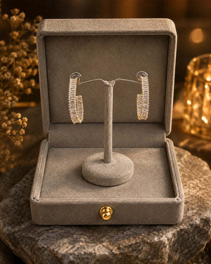 Gold-tone baguette crystal hoop earrings presented on a premium velvet jewelry stand inside a luxury grey jewelry box with soft golden bokeh lighting.