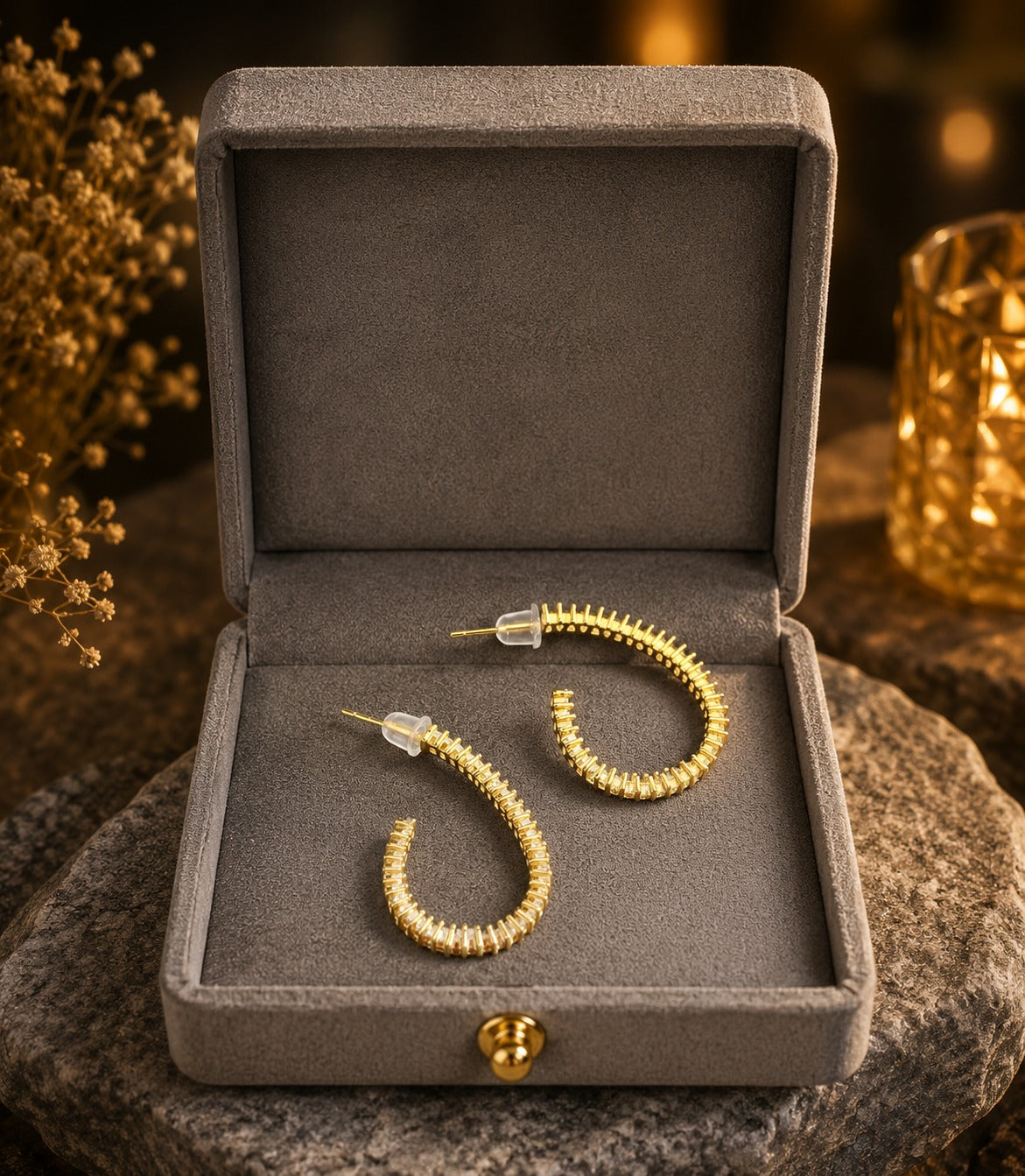 Pair of gold-tone baguette crystal hoop earrings displayed inside a premium grey velvet jewelry box on a natural stone surface with warm ambient lighting.