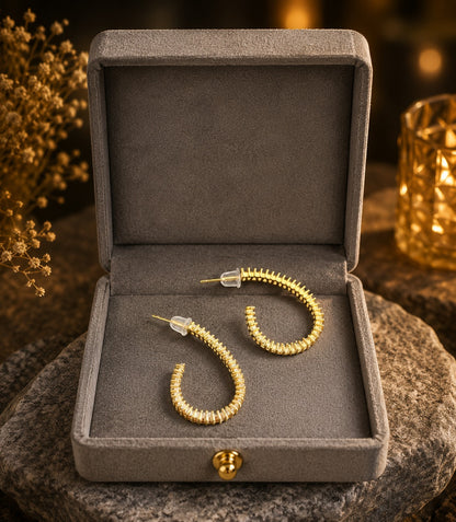 Pair of gold-tone baguette crystal hoop earrings displayed inside a premium grey velvet jewelry box on a natural stone surface with warm ambient lighting.