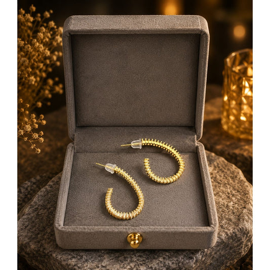 Pair of gold-tone baguette crystal hoop earrings displayed inside a premium grey velvet jewelry box on a natural stone surface with warm ambient lighting.