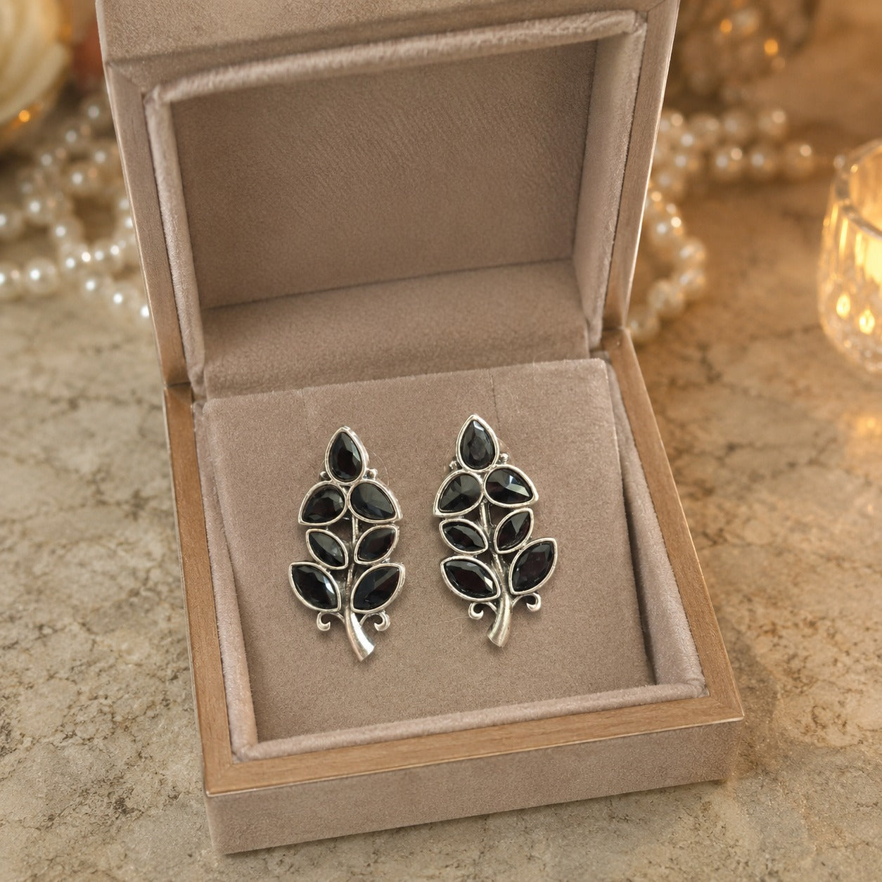 Pair of leaf-shaped earrings with black stones in a jewelry box on a textured surface.