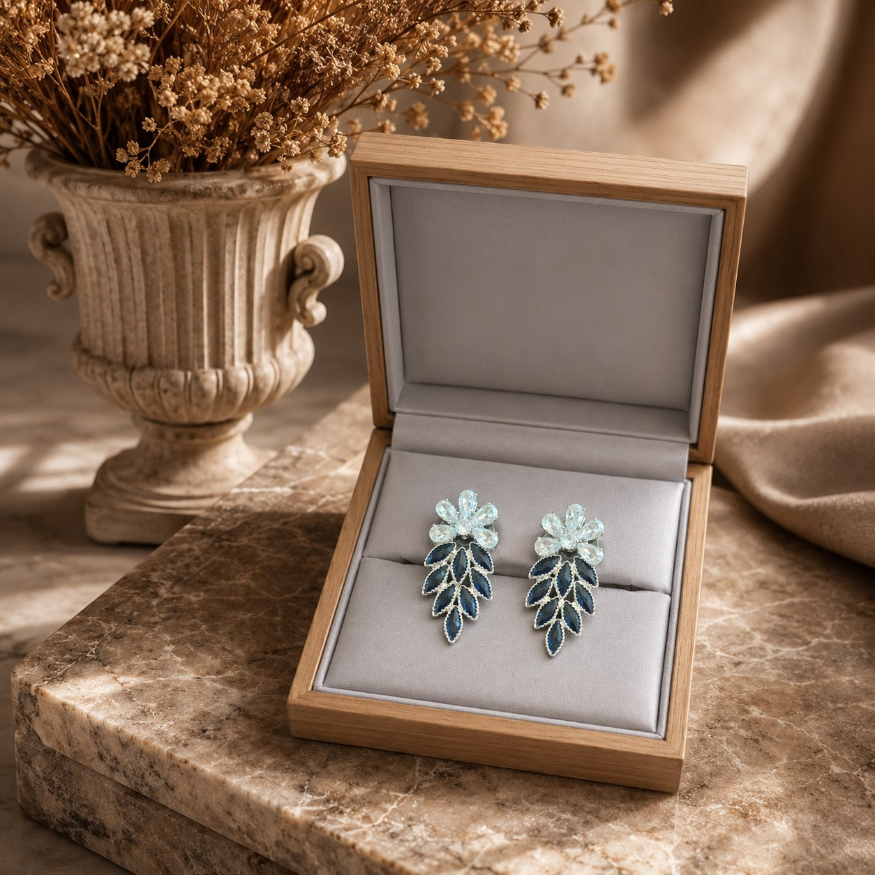 Jewelry box with a pair of leaf-shaped earrings on a marble surface with a vase of dried flowers in the background.