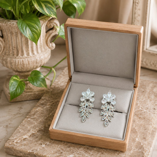 Pair of silver earrings in an open wooden jewelry box on a marble surface with a plant in the background.