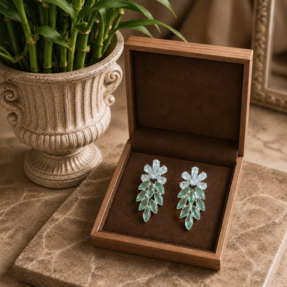 Pair of green leaf-shaped earrings in an open brown jewelry box on a marble surface with a plant in the background.