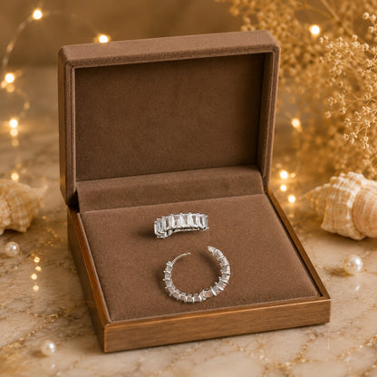 Silver baguette crystal huggie hoop earrings displayed in a premium velvet jewelry box with warm fairy lights and decorative shells.