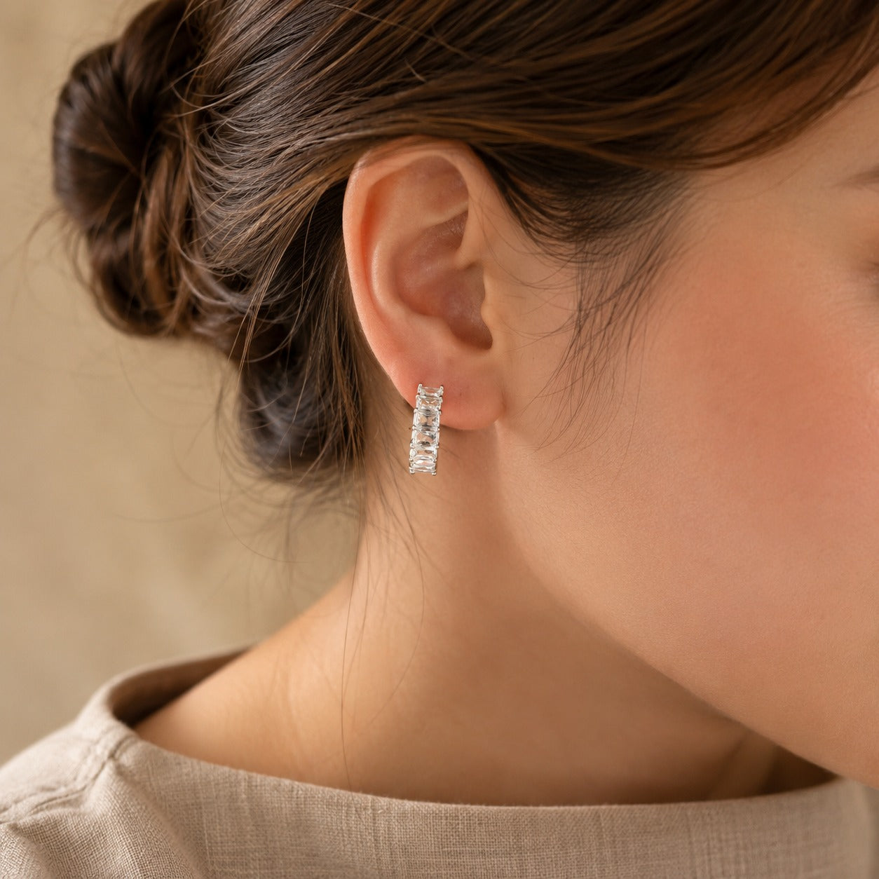 Woman wearing silver-tone baguette crystal huggie hoop earrings in a side-profile portrait with a neutral linen outfit.