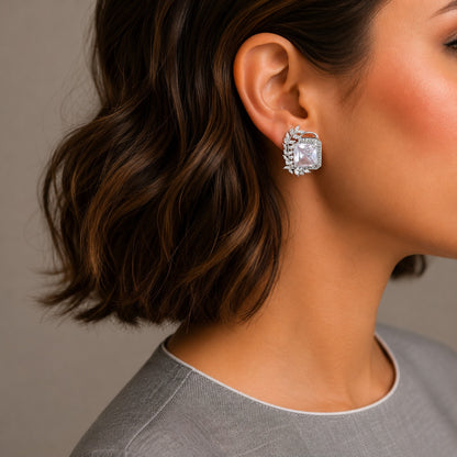 A close-up side profile of a woman wearing a silver-toned square stud earring with a clear faceted gemstone, set against a neutral gray background.
