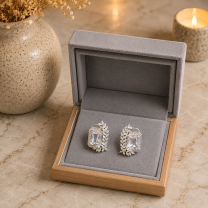 A pair of silver-toned rectangular stud earrings with clear gemstones, presented in an open gray velvet jewelry box on a beige marble surface, with a ceramic vase and a lit candle in the background.