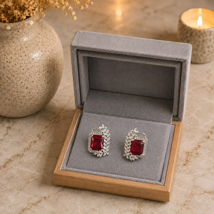 A pair of silver-toned earrings with rectangular red gemstones and small clear stones, displayed in an open gray jewelry box on a marble surface with a vase and lit candle in the background.