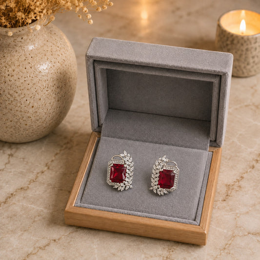 A pair of silver-toned earrings with rectangular red gemstones and small clear stones, displayed in an open gray jewelry box on a marble surface with a vase and lit candle in the background.
