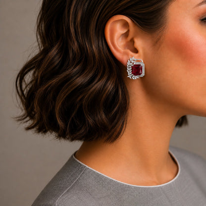 A close-up side profile of a woman with short wavy brown hair wearing a single square-shaped silver-toned stud earring with a deep red center stone and a halo of small clear stones.