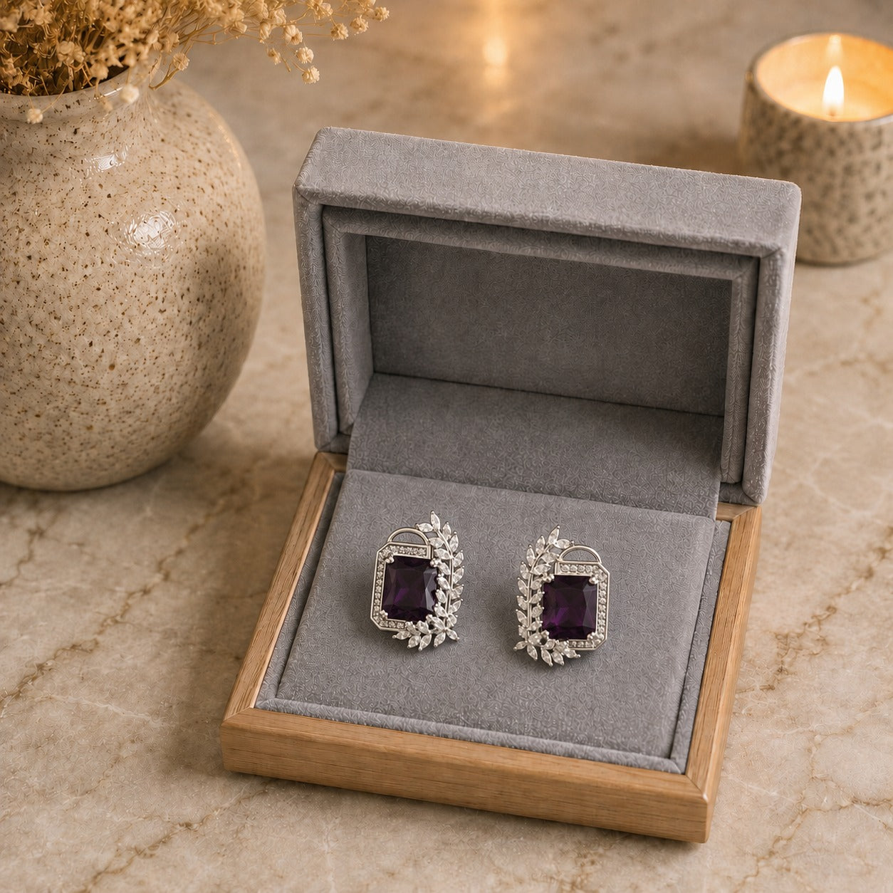 A pair of silver-toned rectangular stud earrings with deep purple rectangular centers and small clear stones, displayed in an open gray jewelry box on a beige stone surface with a vase and lit candle in the background.
