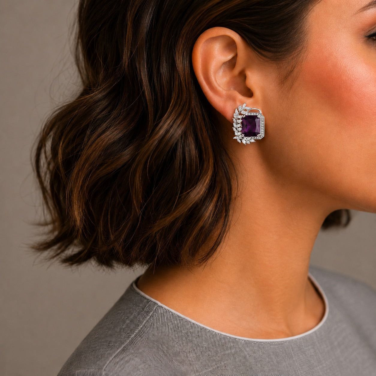 A close-up side profile of a woman with short brown hair wearing a silver-toned square stud earring with a deep purple rectangular center stone and a halo of small clear stones, against a neutral gray background.